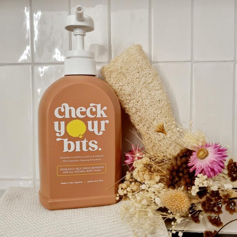 Check Your Bits body wash on a tiled bathroom shelf with a loofah and dried flowers